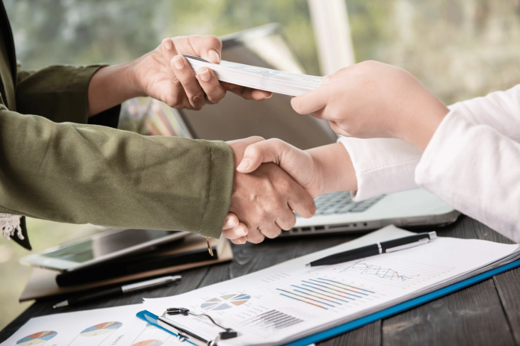 Two people shaking hands at a desk with papers and a laptop.