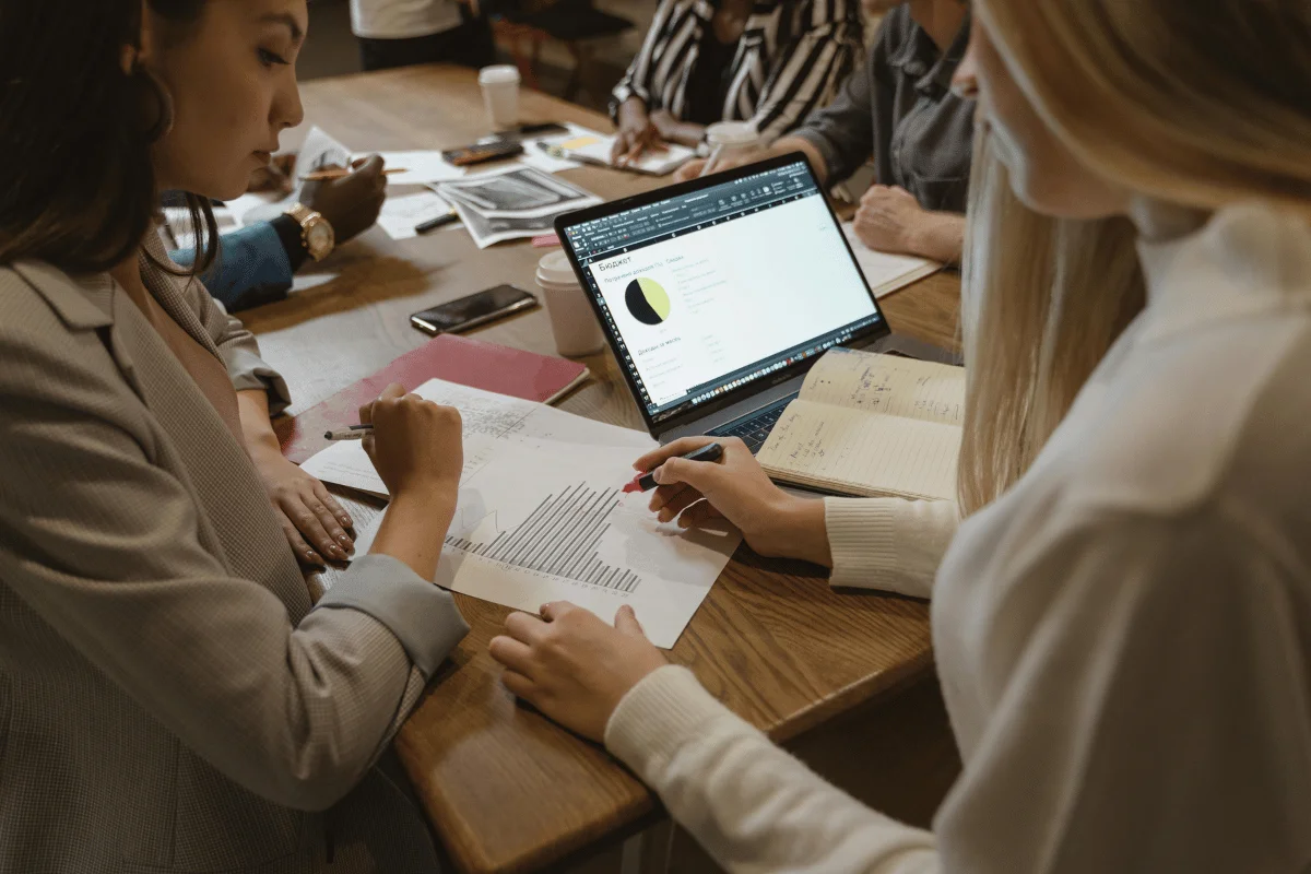Two women analyzing graphs on paper and a laptop screen during a business meeting, with other colleagues in the background.