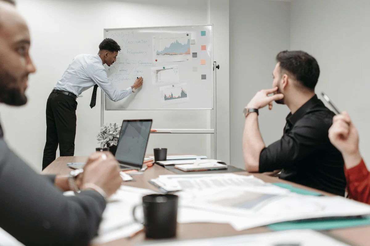 A man in a suit presents data on a whiteboard to a group of colleagues during a business meeting.