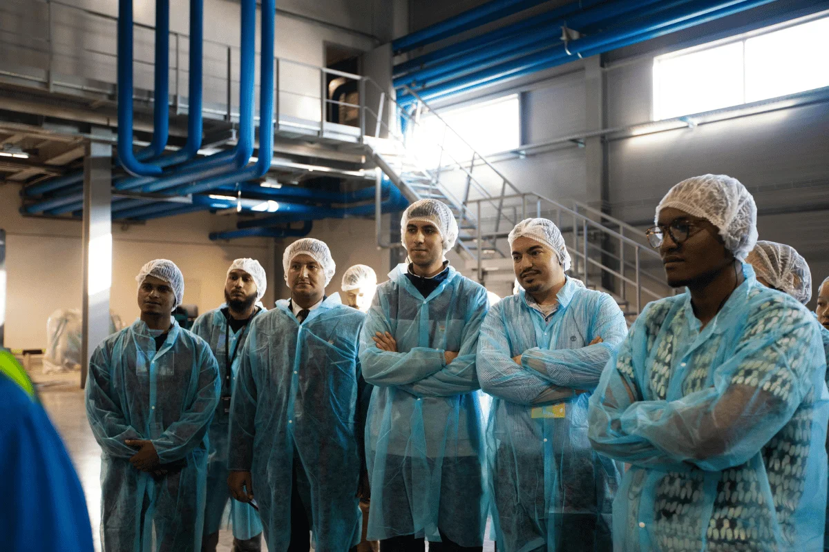 Employees in blue safety coats and hair covers listening to instructions during a factory floor meeting.