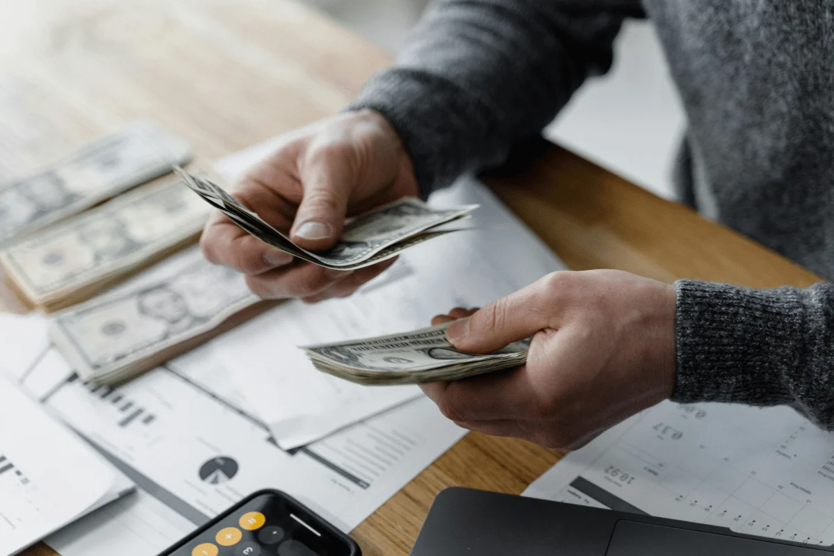 A person counting bills of cash on a desk with financial documents and a calculator.