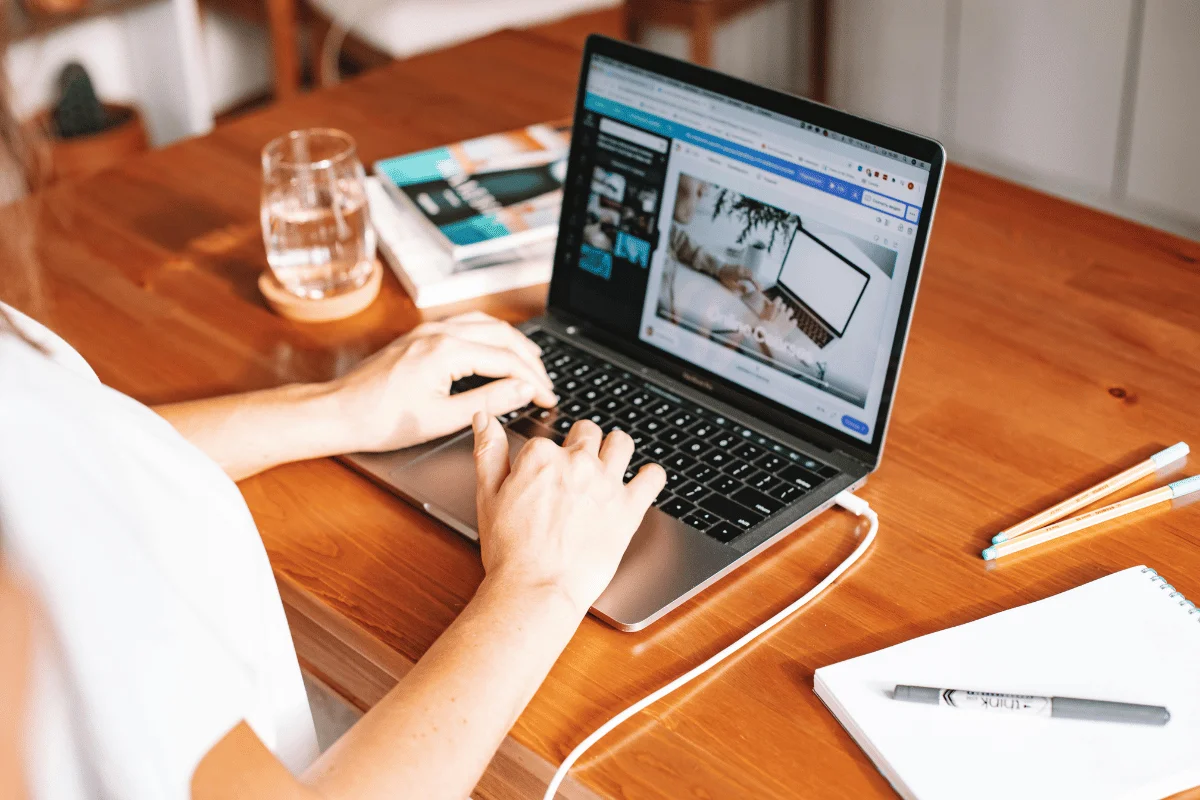 A person is working on a laptop at a wooden table, viewing an image of a workspace with a laptop and notebook.