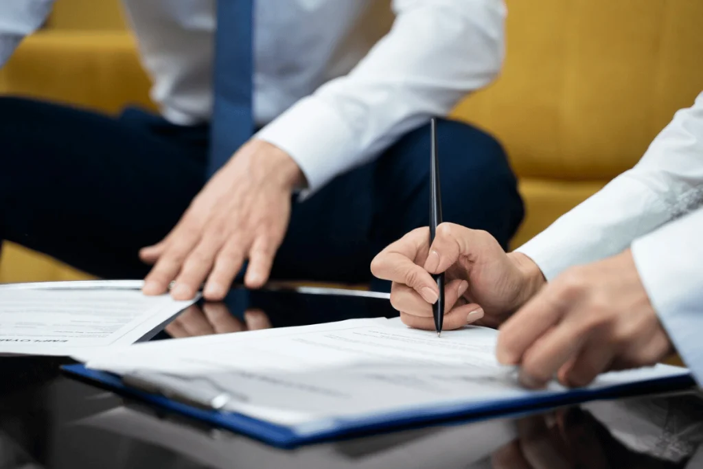 Two individuals in formal attire, one writing on documents while the other rests their hand on the table, engaged in a discussion.