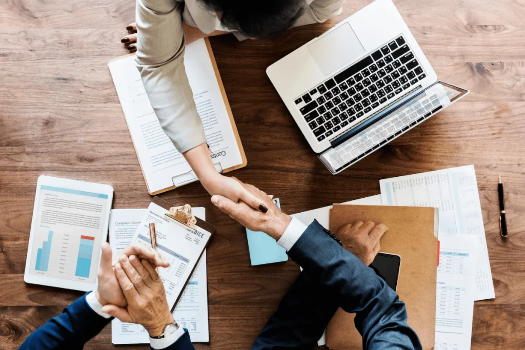 Business people shaking hands at a table with papers and laptops.
