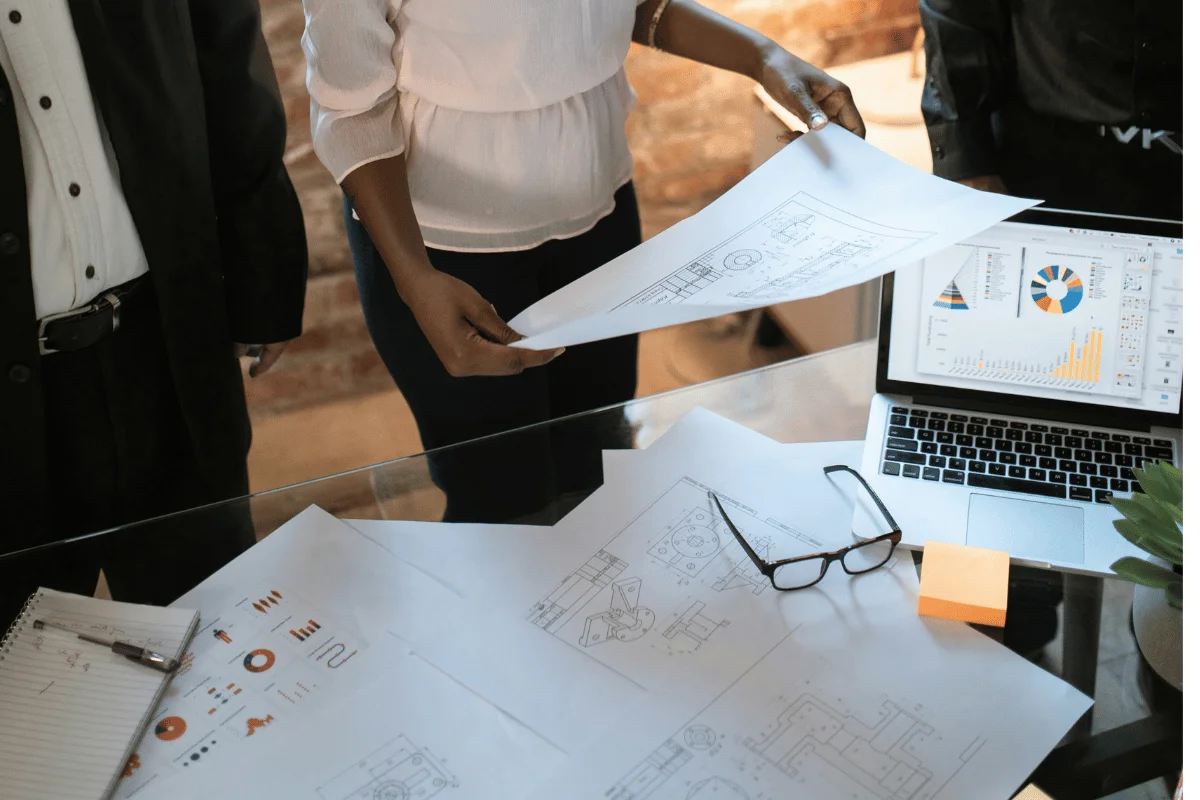 A team examines architectural plans at a glass table, with charts on a laptop and scattered papers. The atmosphere is focused and collaborative.