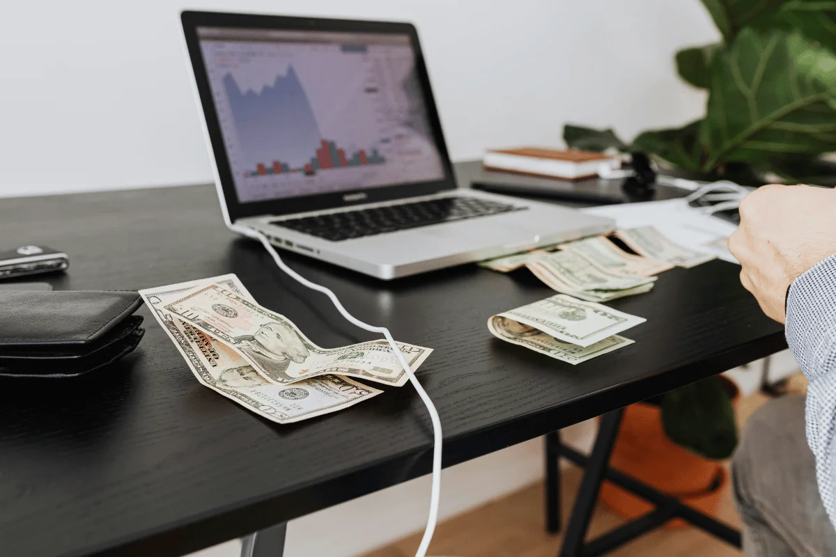 A desk with a laptop showing stock graphs, several bills of cash, and a wallet.