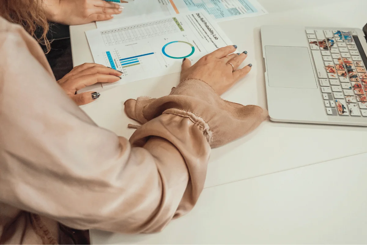 A close-up of a person reviewing data and charts printed on paper while sitting next to a laptop.
