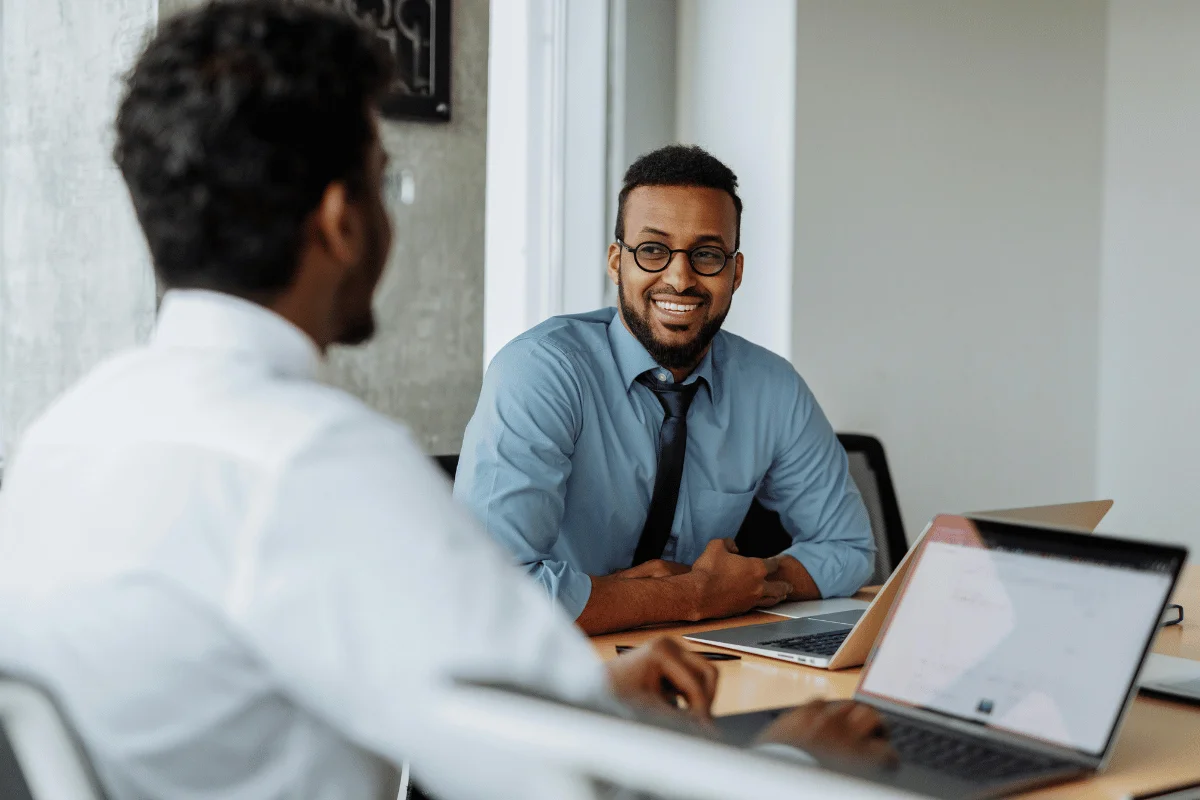 Two business professionals are having a friendly conversation in a modern office, both seated with laptops on the table.
