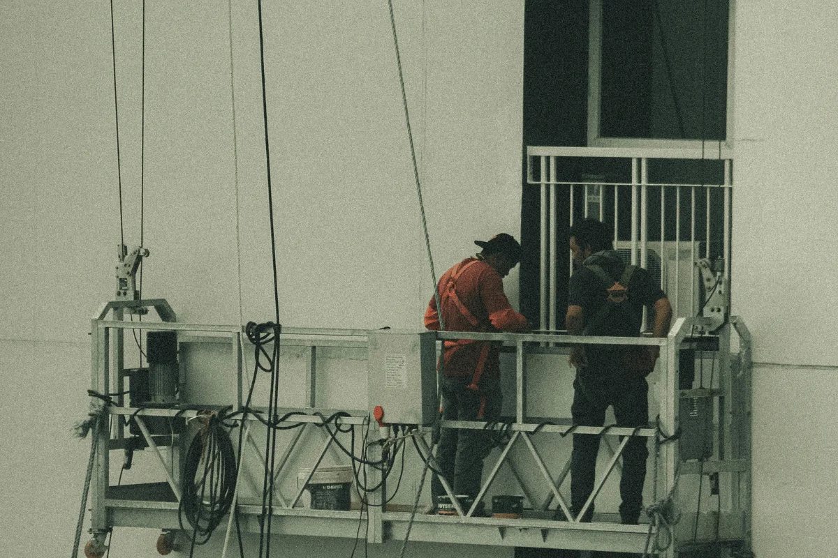 Two workers on a suspended platform in front of a building facade discuss their task. 