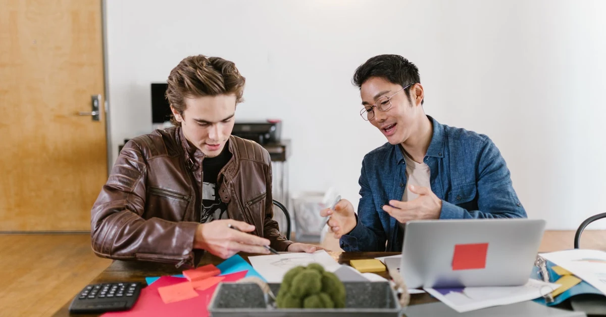 Two young technology business brokers reviewing documents and laptop at wooden desk with colorful sticky notes and calculator.