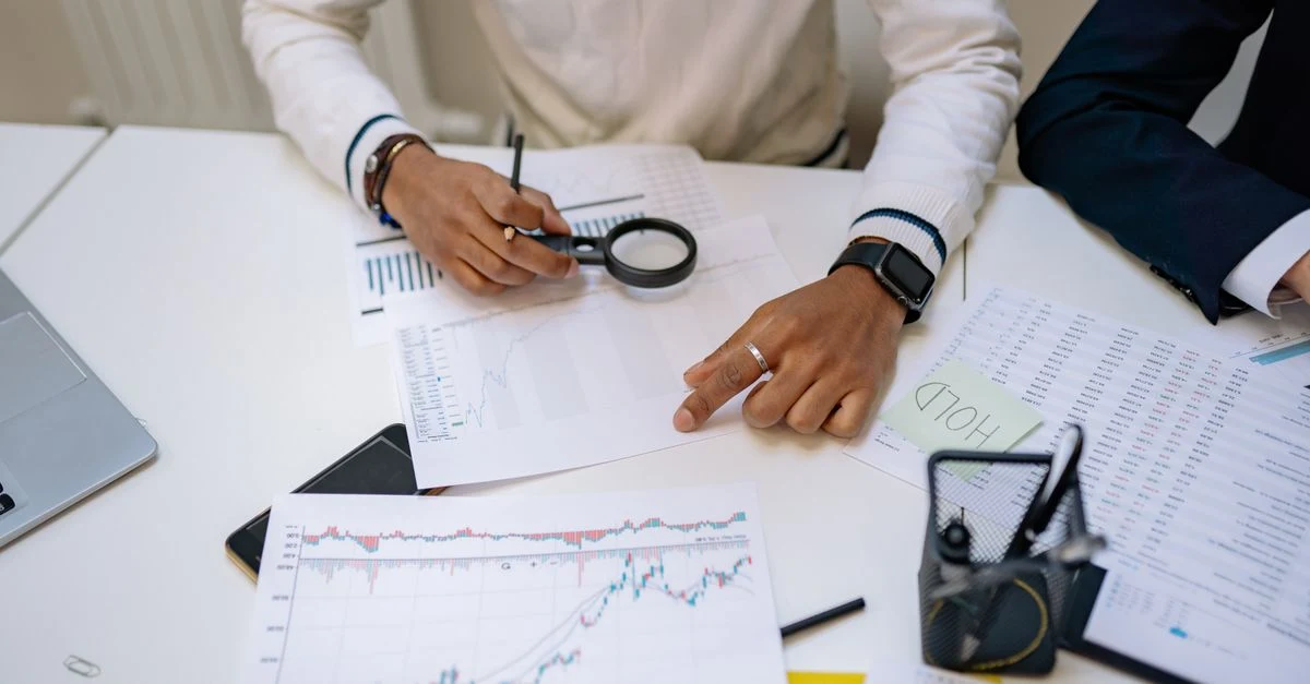 Business professional analyzing stock charts and financial data with magnifying glass on desk.