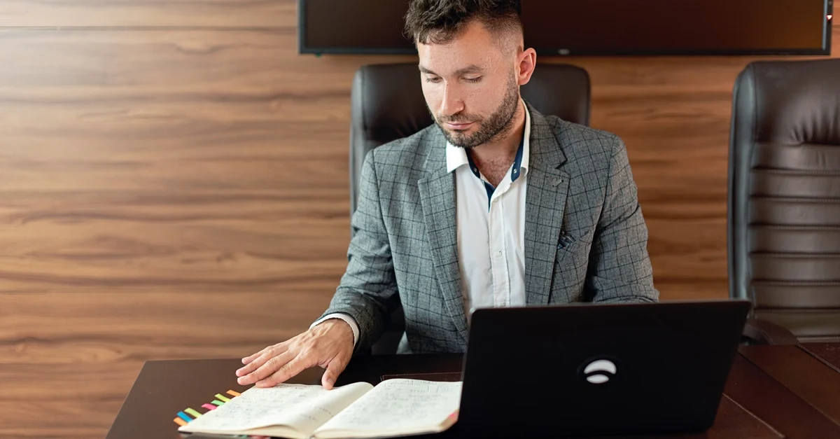 Professional man reviewing business documents and laptop at desk, planning SaaS exit strategy in modern office.
