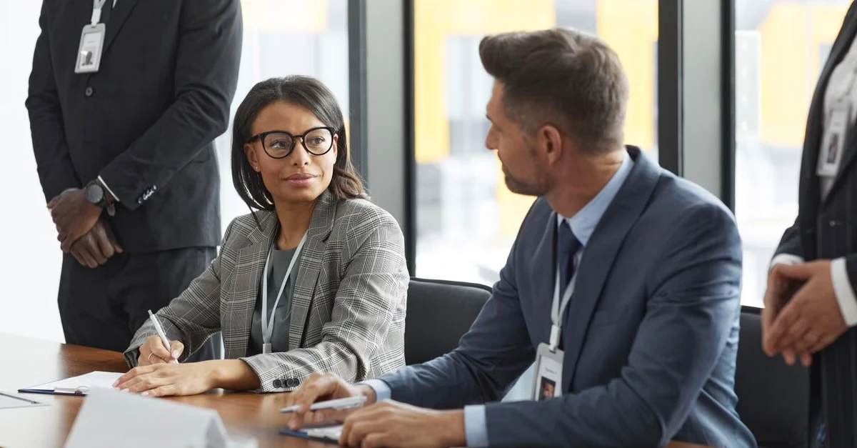 Business professionals in suits discussing at conference table with city skyline behind during agency meeting negotiation.