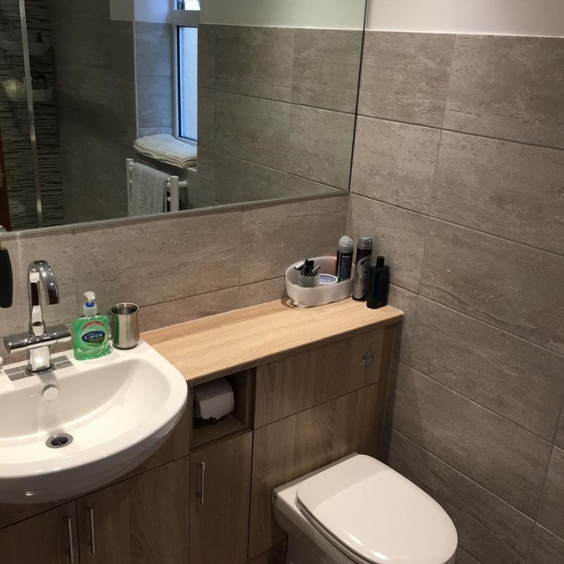 Modern bathroom corner with white sink, chrome faucet, wood cabinet, toilet, and a large mirror reflecting towels on a rack.