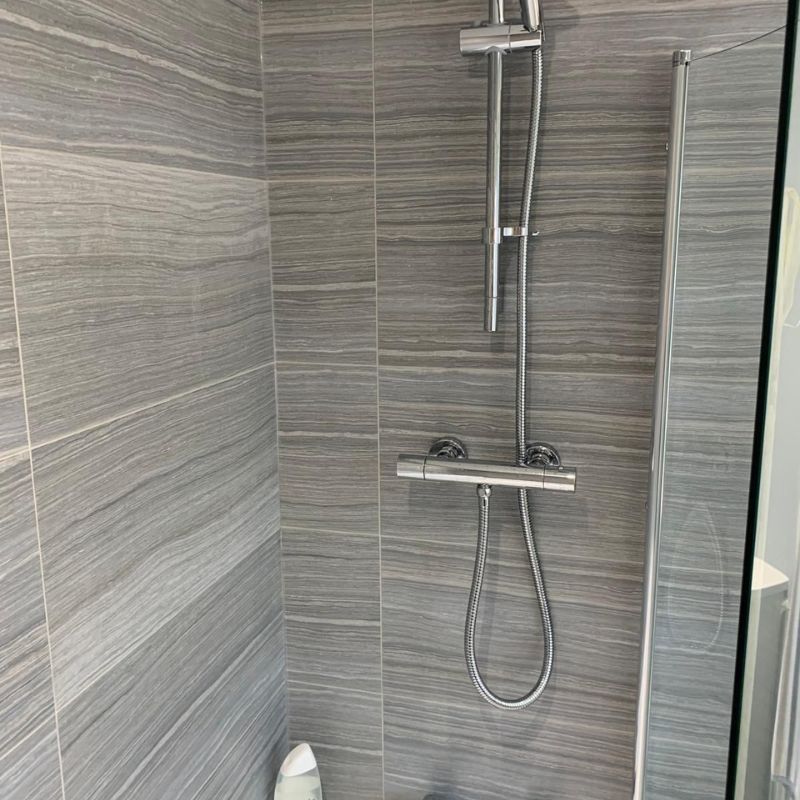 Modern shower area with gray horizontal striped tile walls, a chrome showerhead and handheld shower attached to a metal rail, and a bottle of shower gel on the floor corner.