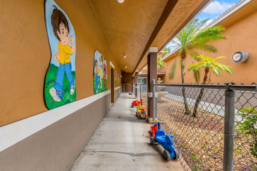 A covered outdoor concrete walkway next to a chain-link fence. The wall is decorated with large, colorful cartoon cutouts of children, and several plastic ride-on toys are parked along the path.