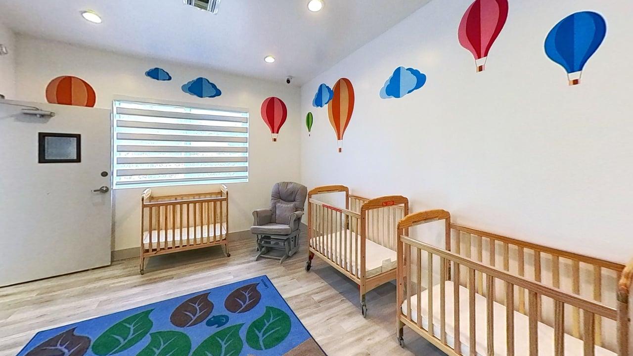 Bright nursery room with four wooden cribs, a gray rocking chair, a window with striped blinds, and colorful hot air balloon and cloud wall decals.