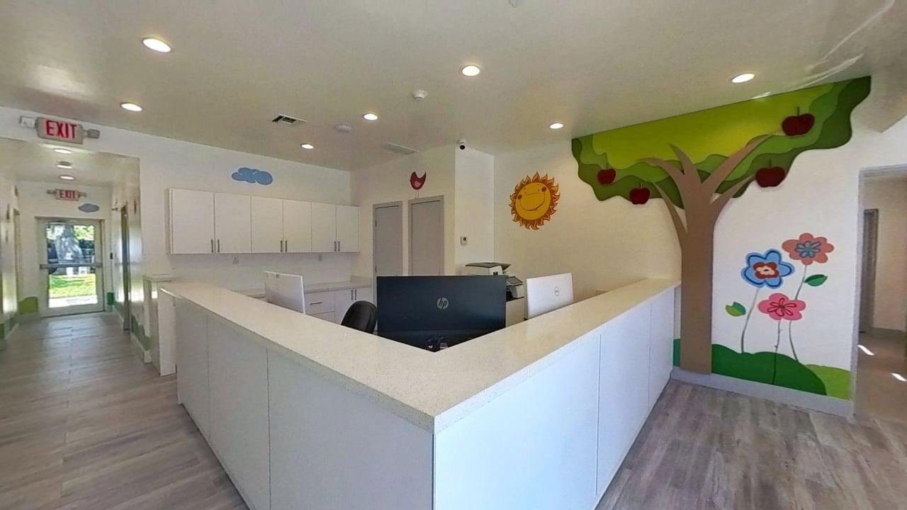 Reception area with white counters, computer monitors, and colorful wall murals of a smiling sun, tree with red apples, and flowers.