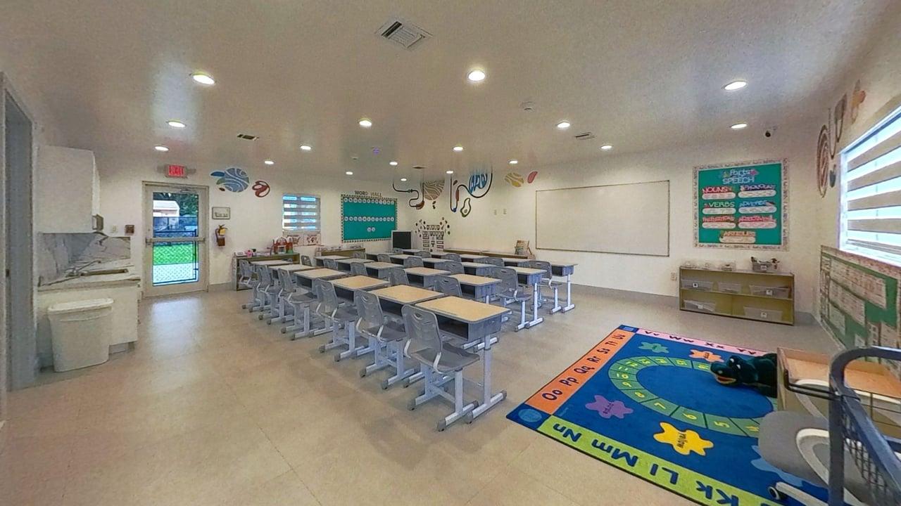 Bright classroom with rows of desks and chairs, colorful alphabet rug, whiteboard, and educational posters on the walls.