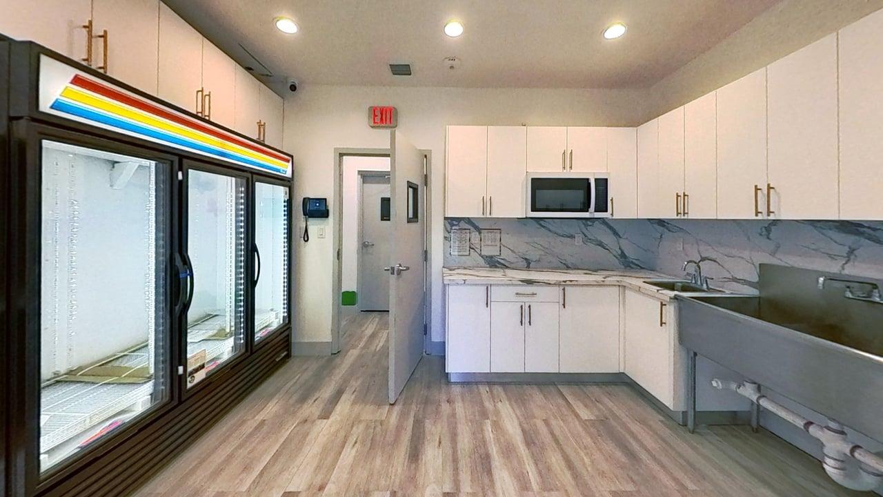 Modern kitchen with white cabinets, marble backsplash, microwave, large stainless steel sink, and empty glass-door refrigerators on the left.