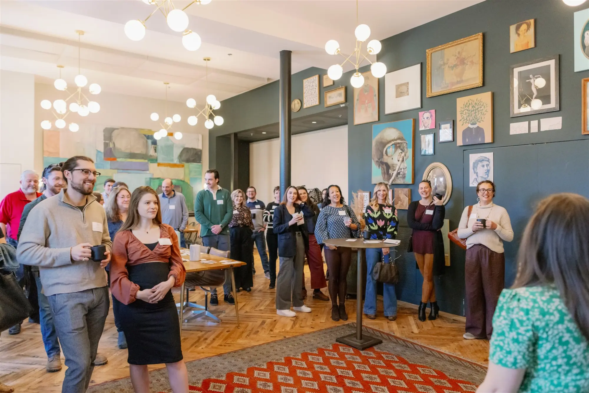 A group of people stand and listen during an indoor gathering with art on the walls.