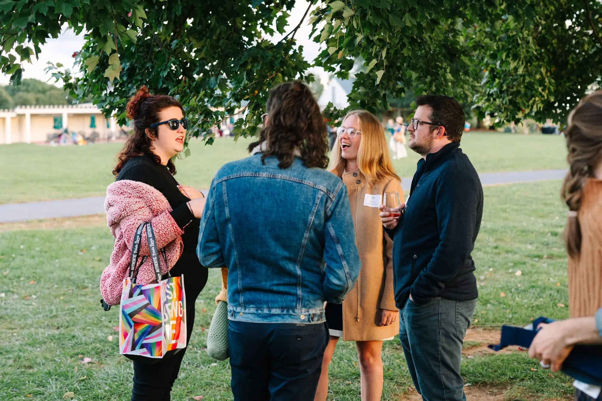 A group of people standing and talking outdoors on a grassy area under a tree.