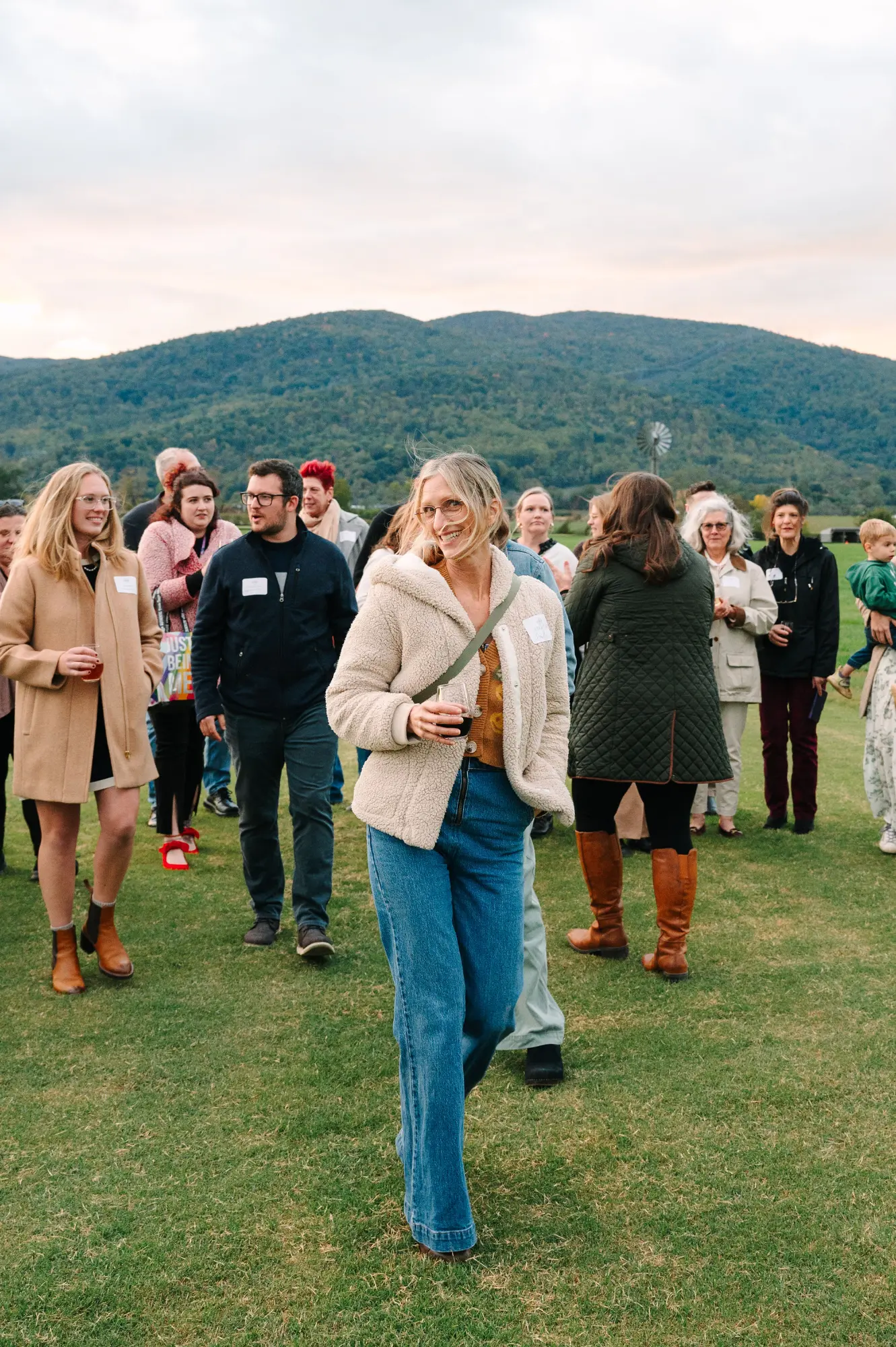 A group of people stands on a grassy field, some holding drinks, near a mountain landscape.