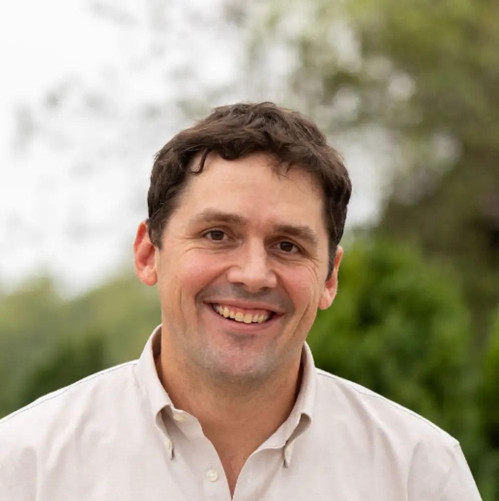 Smiling man with short dark hair wearing a light beige button-up shirt outdoors with blurred greenery in the background.