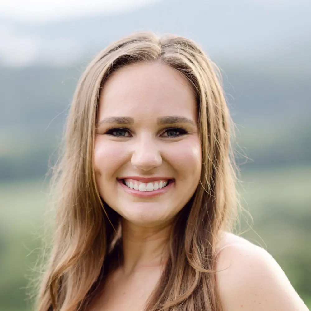 Smiling young woman with long light brown hair in an outdoor setting with a blurred natural background.