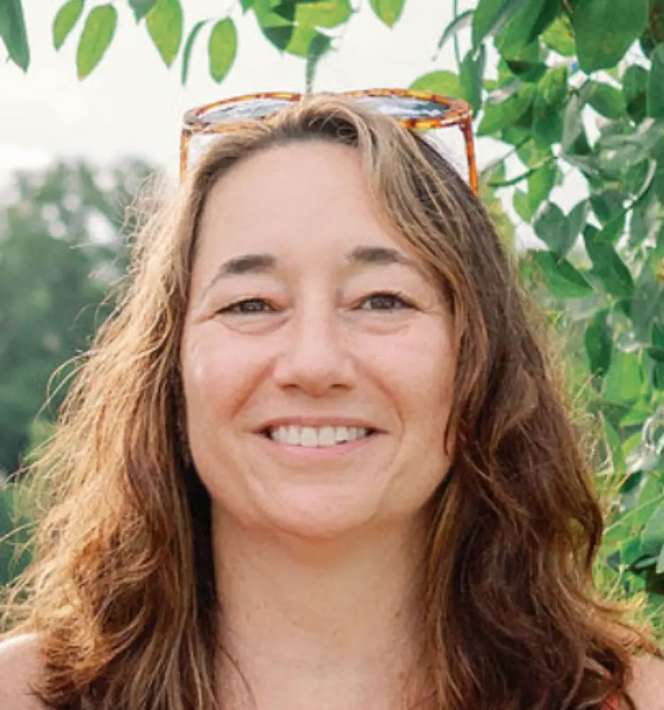 Smiling woman with long brown hair standing outdoors with green leaves in the background and sunglasses on her head.