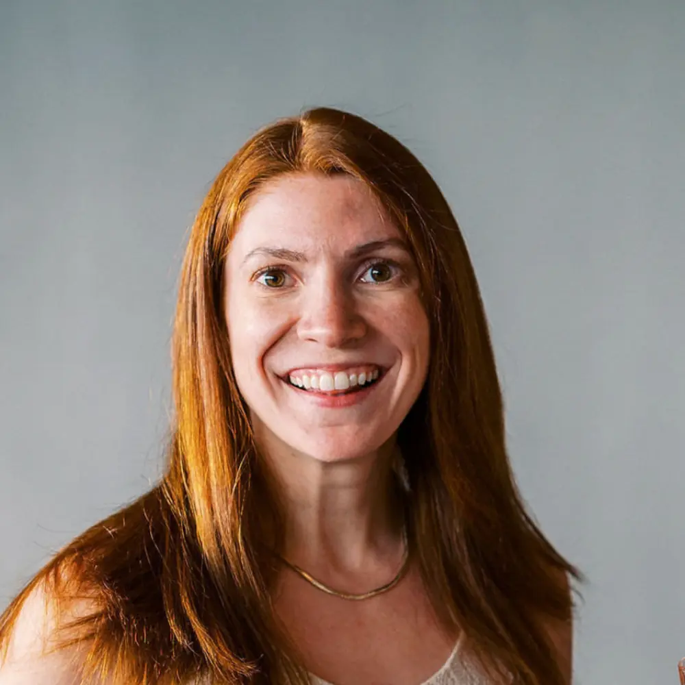 Smiling woman with long red hair wearing a light-colored top and gold necklace against a gray background.