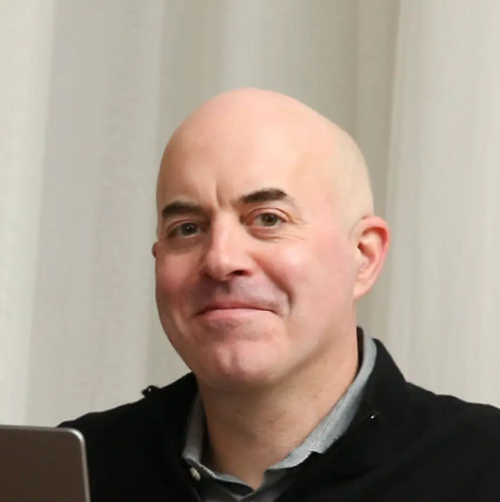 Smiling bald man wearing a black jacket and light gray collared shirt in front of a white curtain.