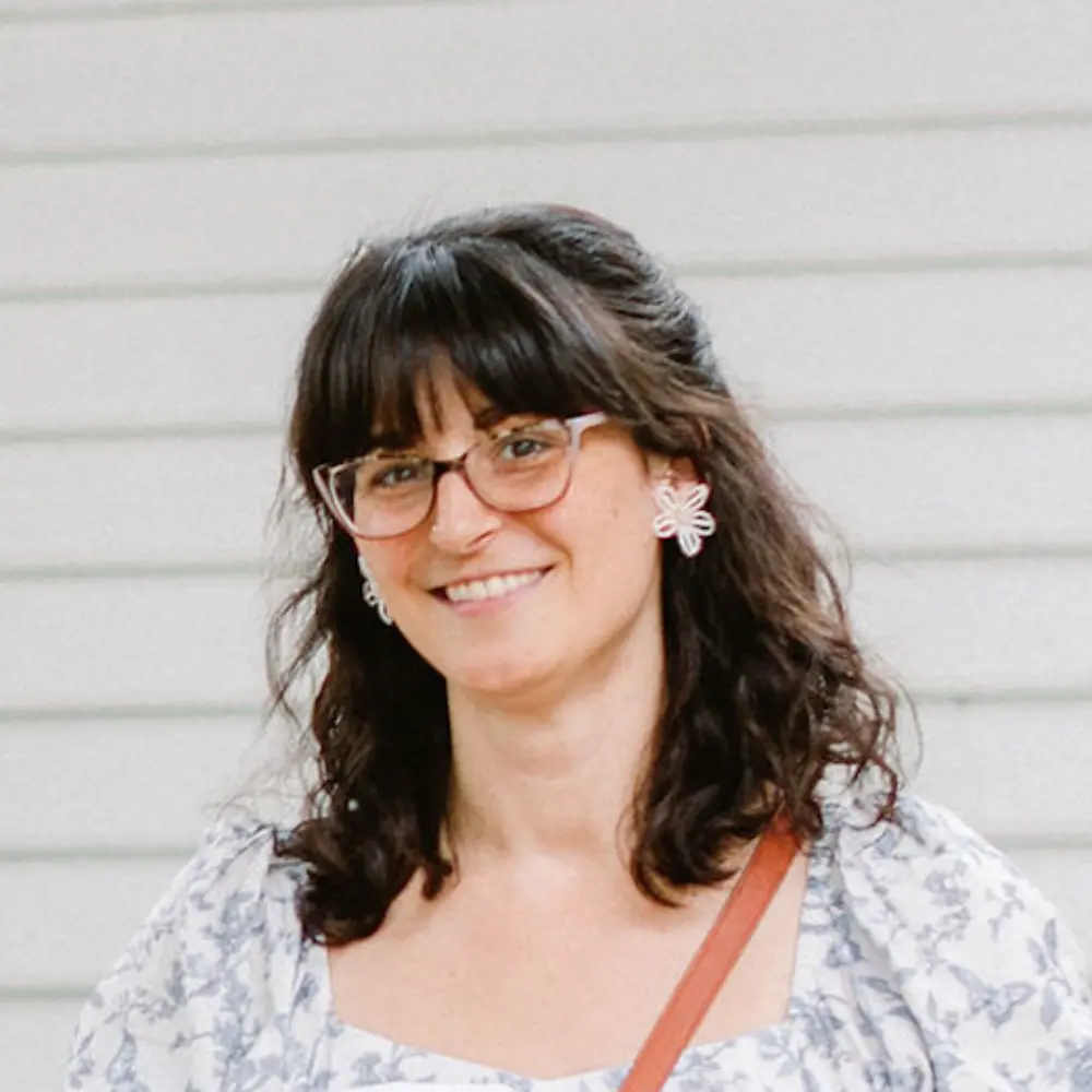 Smiling woman with dark curly hair, wearing glasses, flower-shaped earrings, and a white floral blouse.