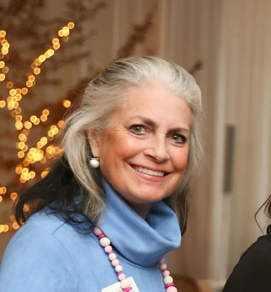 Smiling older woman with gray hair wearing a blue turtleneck and beaded necklace.