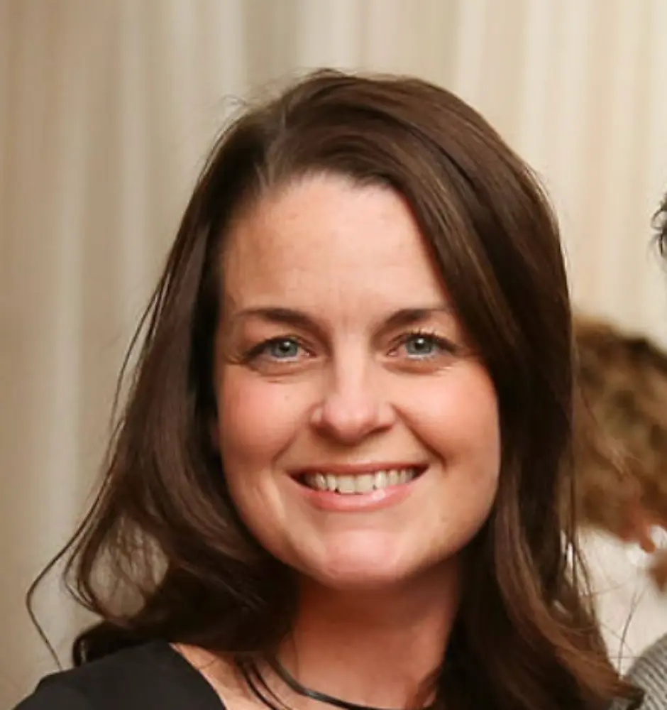 Smiling woman with long dark hair wearing a black top and a black necklace against a neutral background.