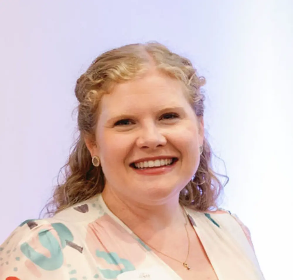Smiling woman with curly blonde hair wearing a patterned blouse and gold necklace against a light background.