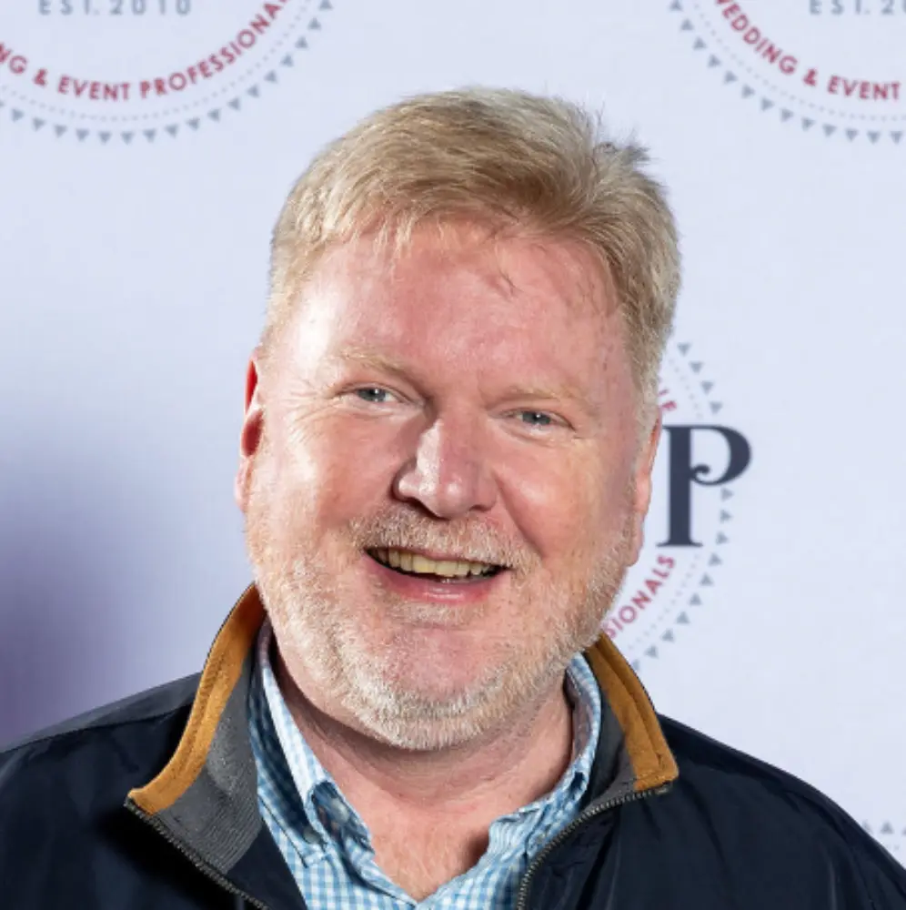Smiling middle-aged man with light hair and beard wearing a checkered shirt and dark jacket.