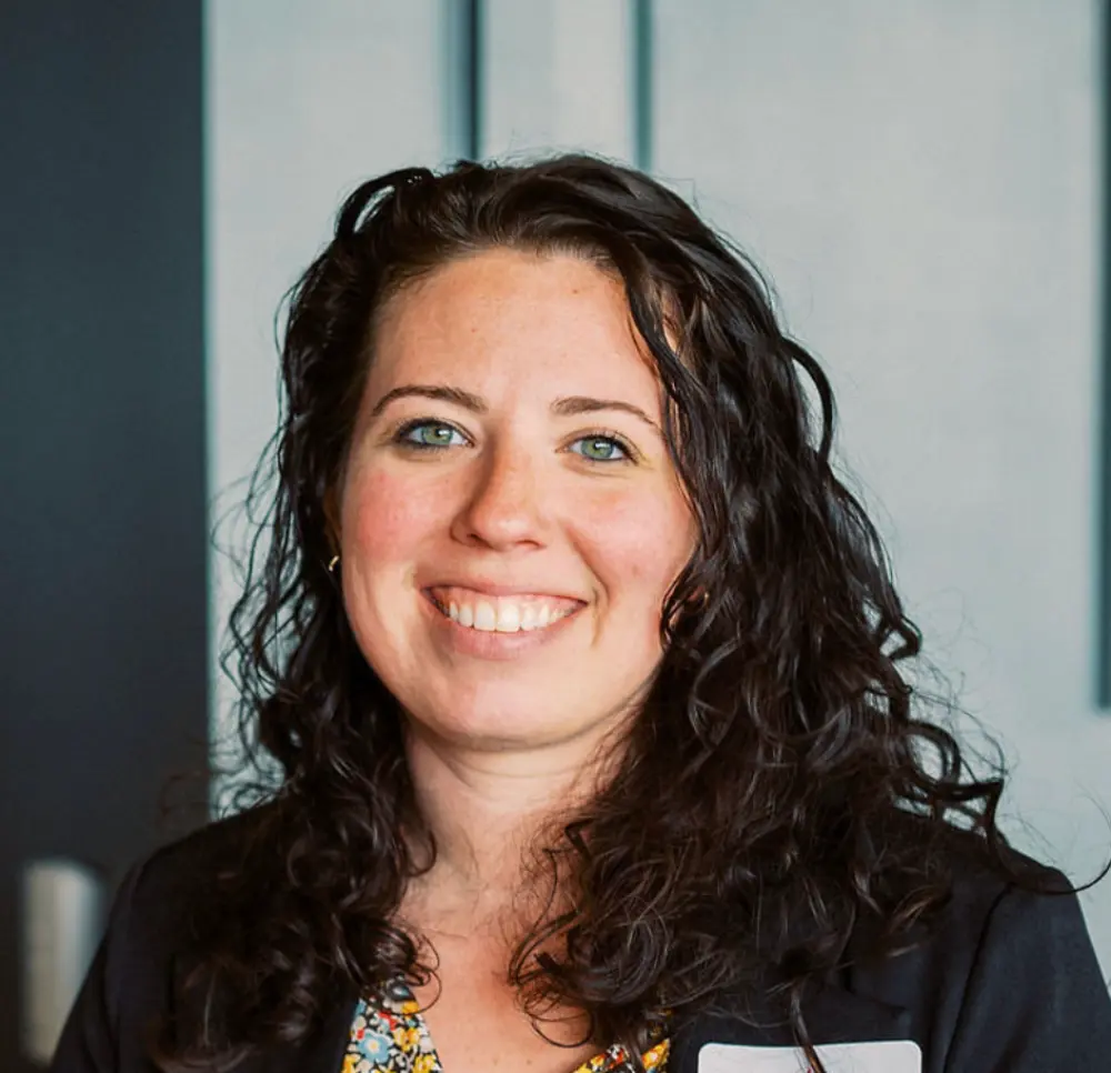 Smiling woman with curly dark hair wearing a black jacket and floral top against a blurred background.