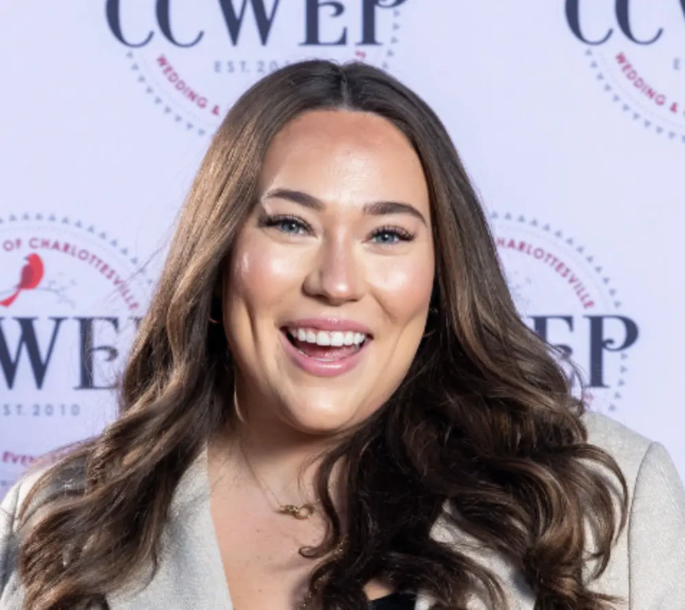 Smiling woman with long brown wavy hair wearing a beige blazer in front of a backdrop with CCWEP logos.