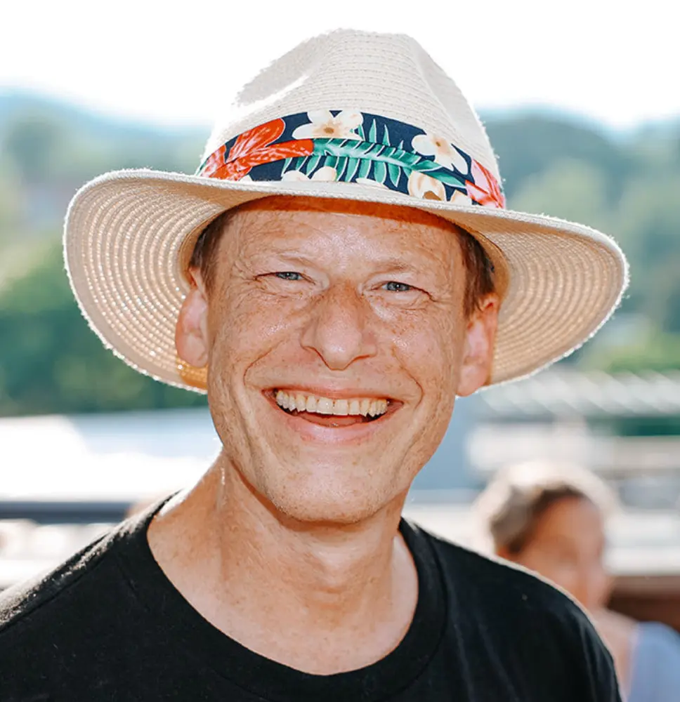 Smiling man wearing a white woven hat with a colorful floral band outdoors.