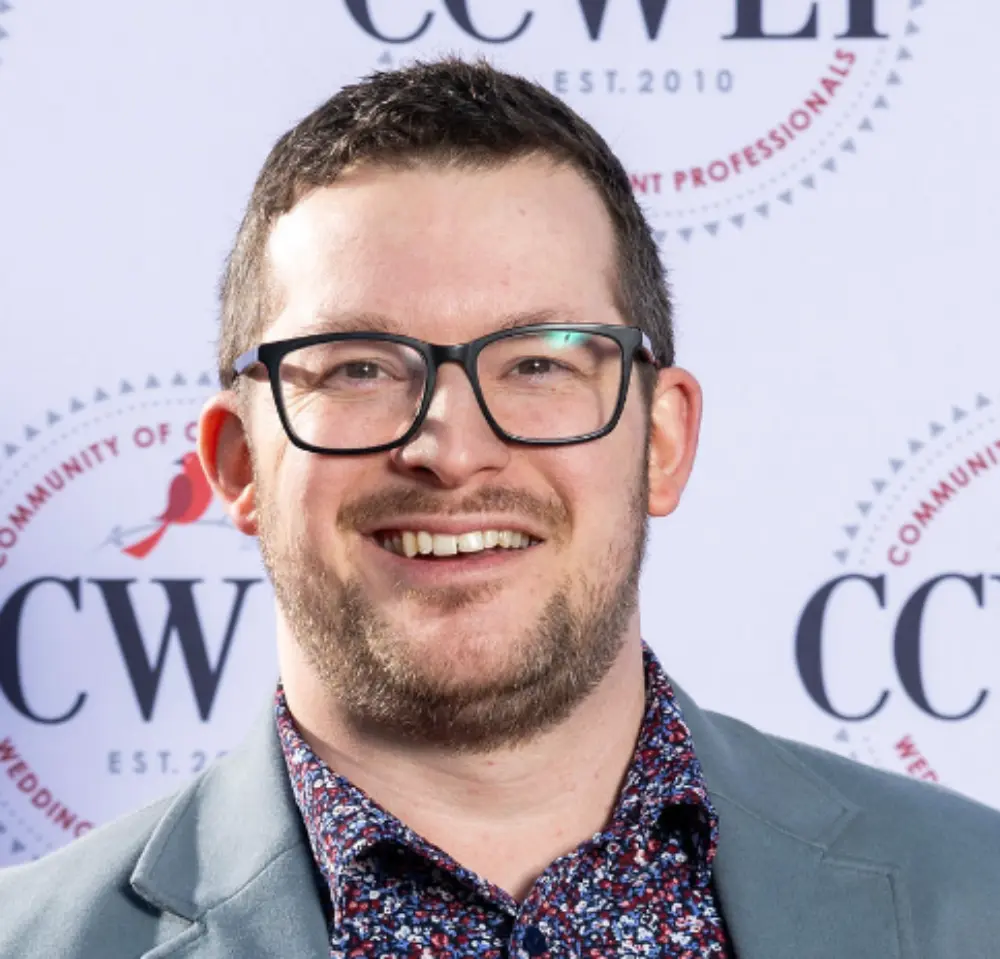 Smiling man with short hair and glasses wearing a patterned shirt and gray blazer in front of a CCWLP backdrop.