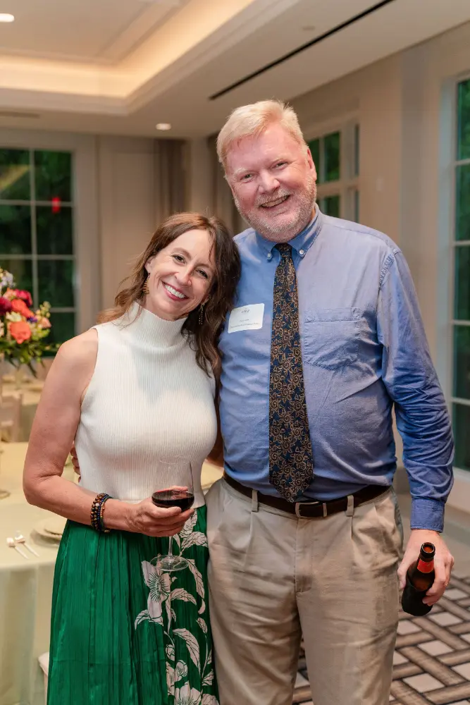 Smiling woman in a white sleeveless top and green skirt and man in blue shirt and patterned tie posing indoors, holding wine and beer bottles.