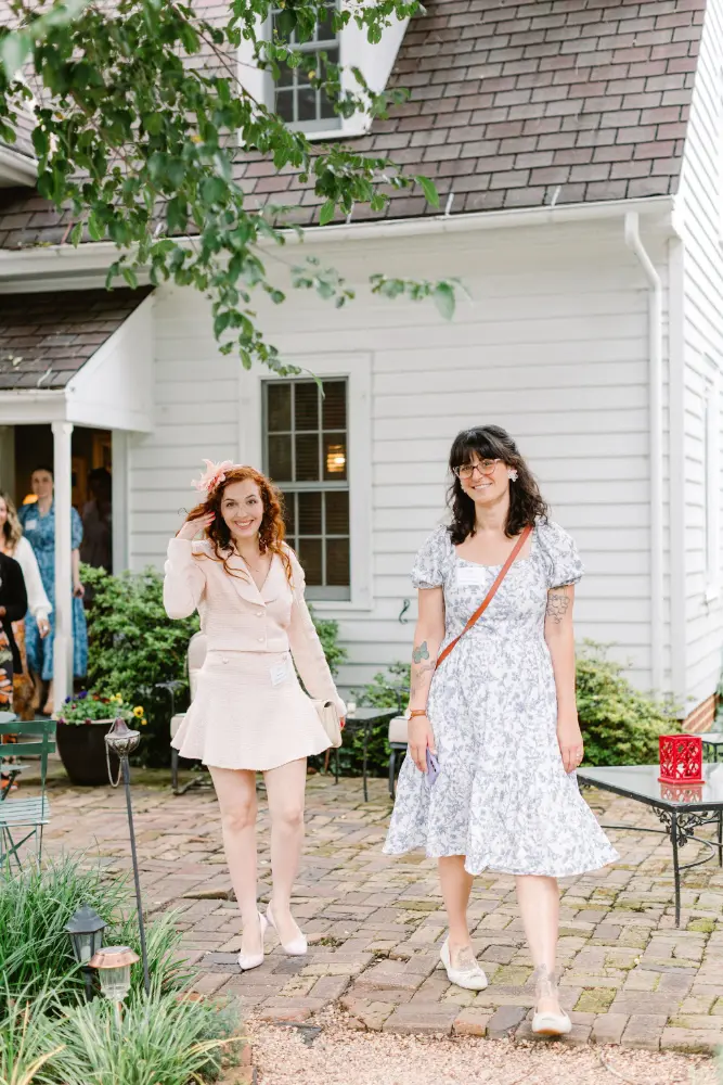 Two women smiling and walking on a brick patio in front of a white house surrounded by greenery.
