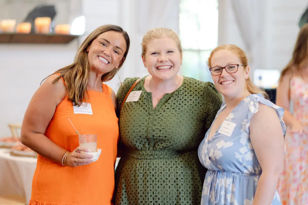 Three smiling women posing indoors, one wearing an orange dress holding a drink, the others in green and blue floral dresses.