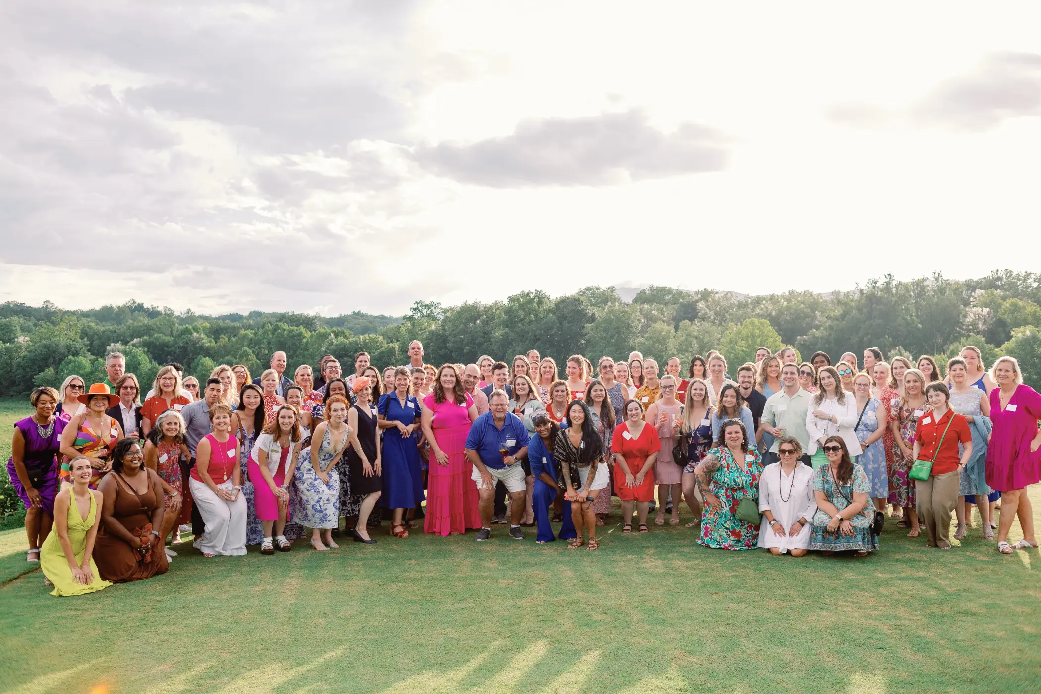 Large group of people posing outdoors on green grass with trees and cloudy sky in the background.