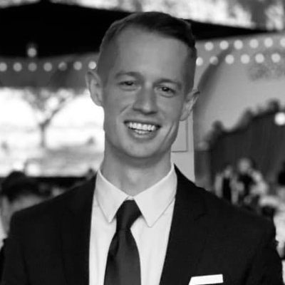 Young man smiling, wearing a black suit and tie with a white shirt and pocket square in an indoor setting with string lights in the background.