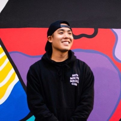 Smiling young man wearing a black backwards cap and black hoodie with 'Anti Social Social Club' text, standing in front of a colorful mural.