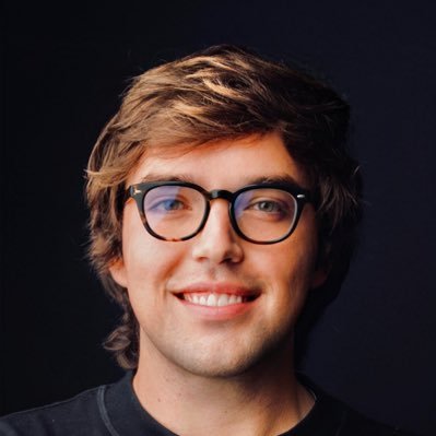 Young man with light brown hair and black round glasses smiling against a dark background.