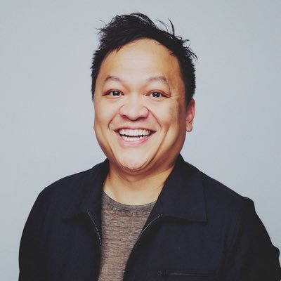 Smiling man with short, dark hair wearing a black jacket over a brown shirt against a plain light background.