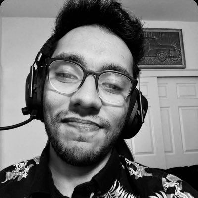 Young man with glasses and headset smiling, wearing a floral patterned shirt in a room with closed doors and a framed artwork behind.